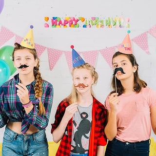 Children wearing colorful pom pom party cone hats with “MAKE” and “PARTY TIME” text