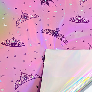 Iridescent pink gift wrap close-up with navy crowns, hearts, and confetti shapes