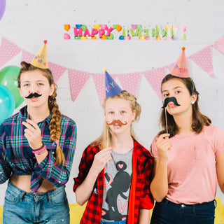 Children wearing pastel rainbow party hats in cone shape with pom pom, printed with phrases like "Make a Wish," "Party Time," and "Yay!!!"