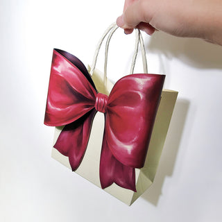 Hand holding cream gift bag with large burgundy bow and rope handles