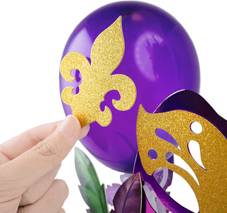 Gold glitter fleur-de-lis mask cutout held over a purple balloon-style topper